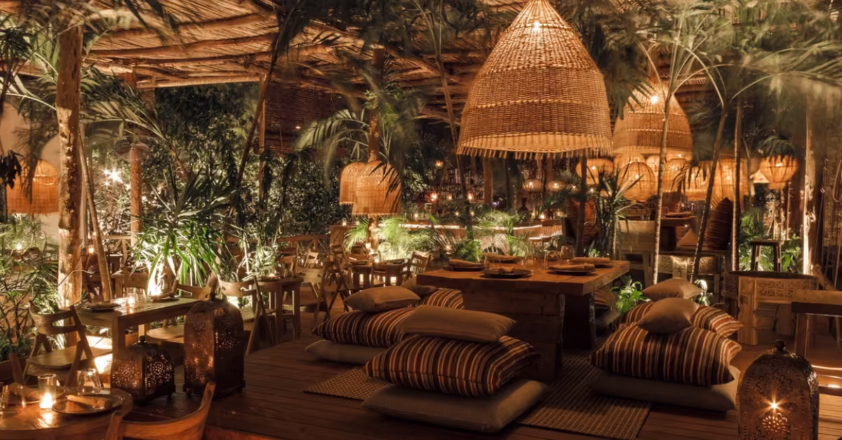 A warm, dimly lit restaurant with woven lamps, lush plants, wooden tables, and floor seating with striped cushions.