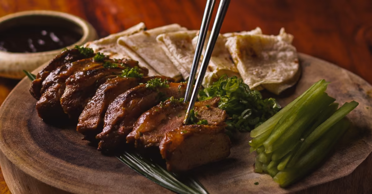 Sliced roasted duck with green onions, cucumber, flatbreads, and dark sauce on a wooden platter; chopsticks lift a piece.