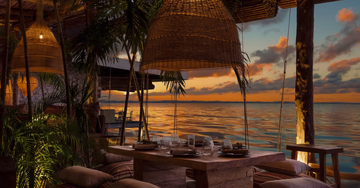 Outdoor dining at Funky Geisha with a rustic table, cushions, wicker lamps, and a sunset view over calm water in orange-purple hues.