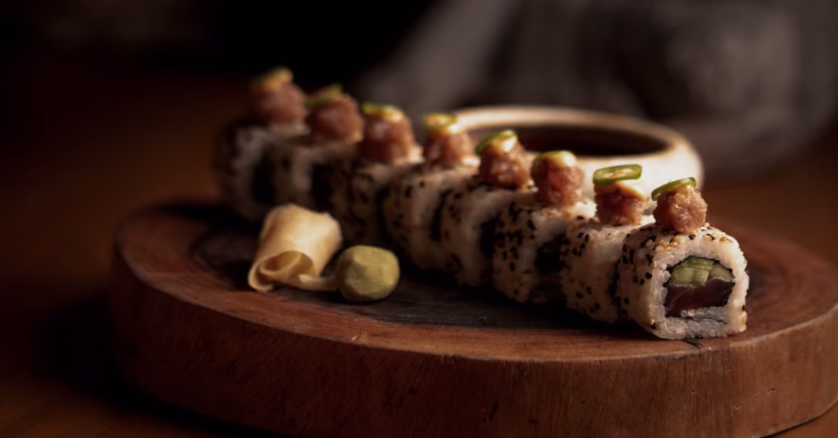 Sushi roll topped with spicy tuna and avocado, served on a wooden board with wasabi, pickled ginger, and soy sauce.