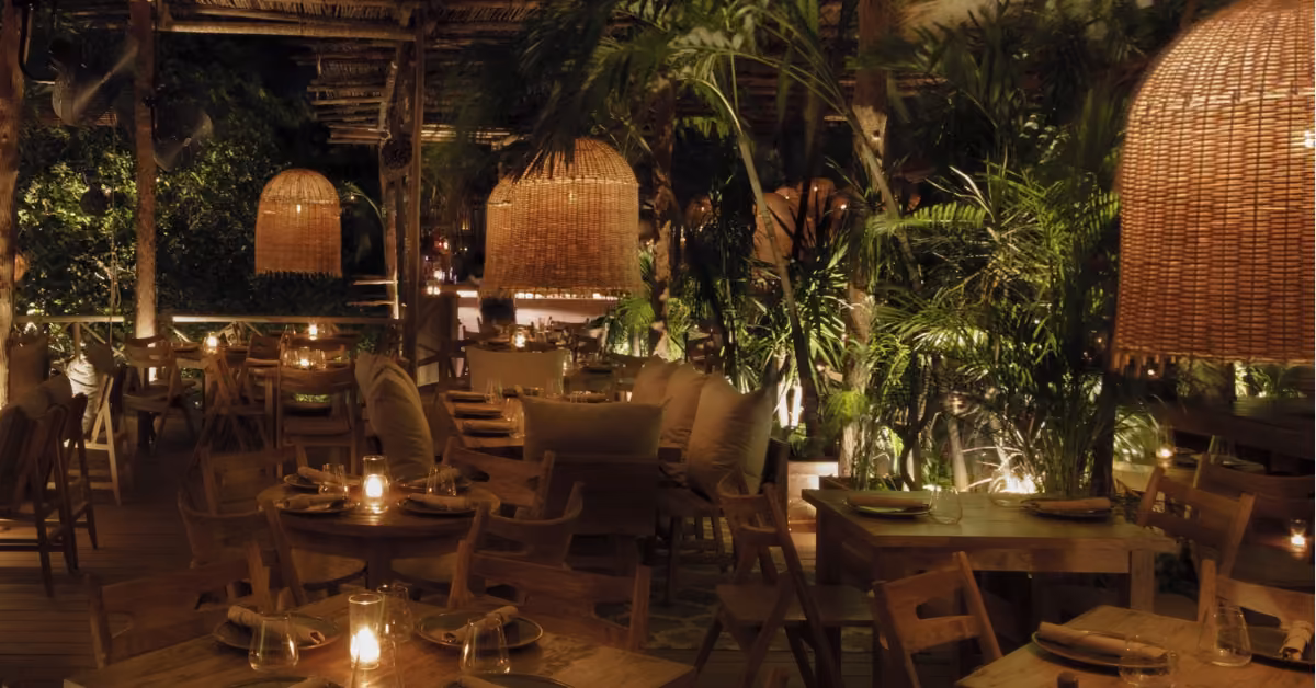 Cozy outdoor restaurant at night with wooden tables, rattan lights, green plants, and candles creating a warm, inviting vibe.