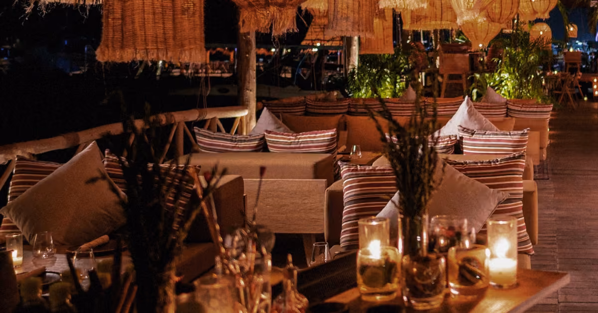 Cozy outdoor lounge at night with striped pillows, wicker lamps, candles, and lush plants for a warm, inviting ambiance.