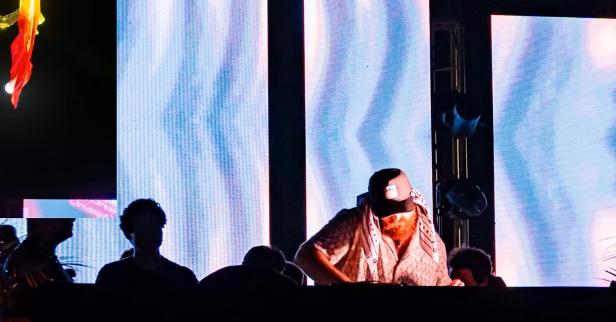 A DJ in a cap and scarf performs at H Roof Night Club, with crowds and vivid blue-purple LED screens in the background.