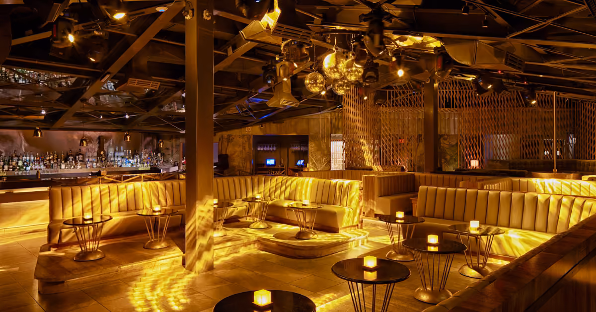 A dim night club with plush curved booths, candlelit tables, a bar in back, gold lighting, and disco balls overhead.