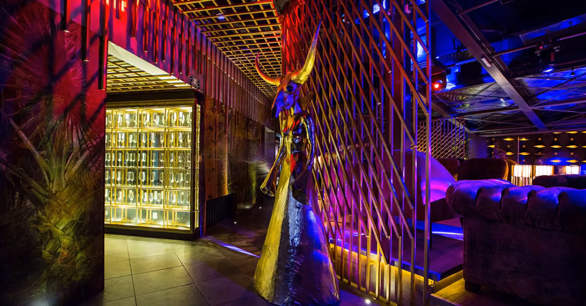 A stylish lounge at H Roof Night Club with colorful lights, plush seating, a sculpture, wooden partitions, and bottle displays.