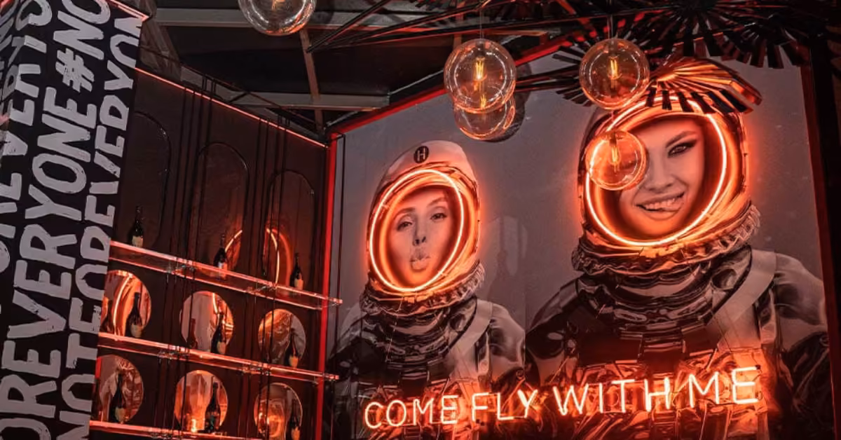 Neon-lit room with two smiling women in spacesuits, a glowing “COME FLY WITH ME” sign, and “FOREVER YOUNG” on the wall.