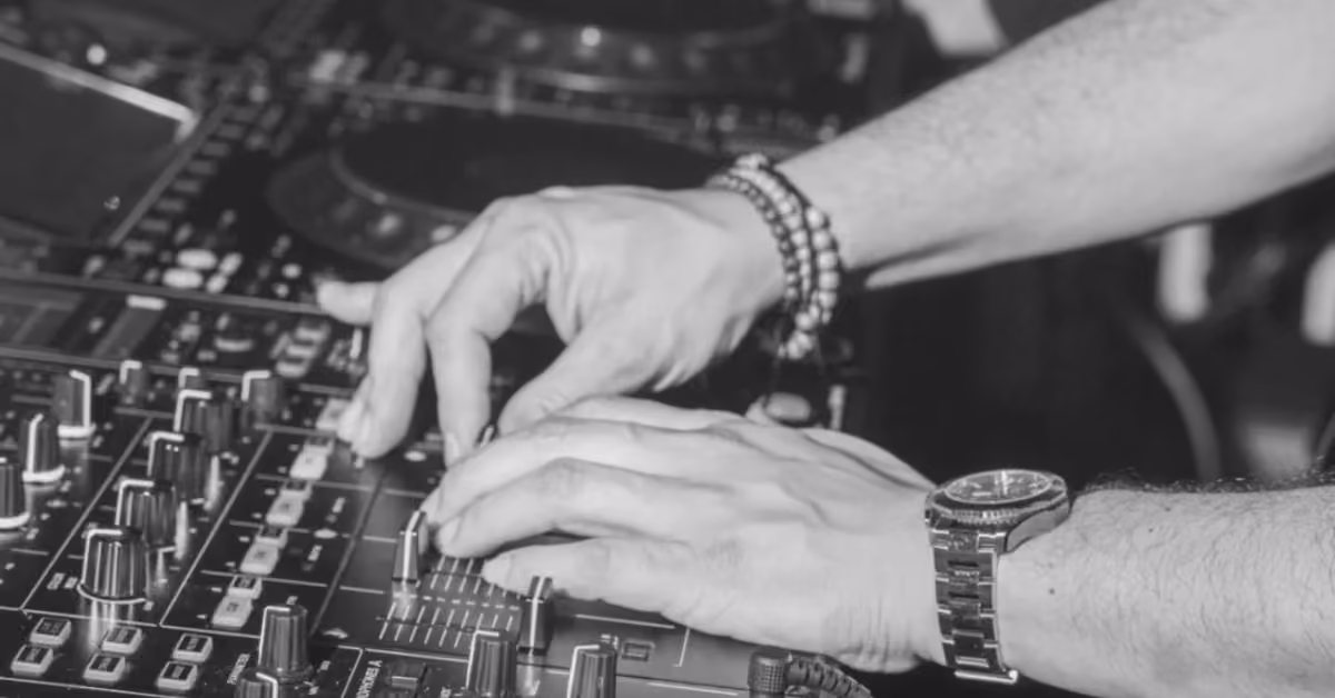 Black and white photo of hands adjusting a DJ mixer at a night club, wearing a watch and bracelets among knobs and sliders.