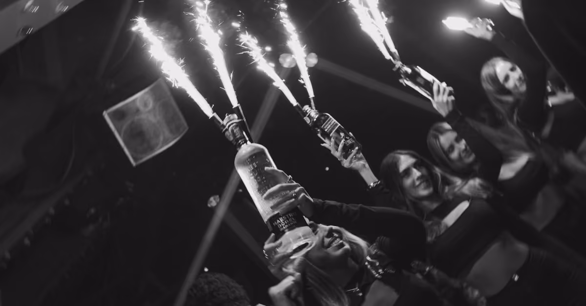 Women celebrate at a rooftop night club, holding bottles with sparklers in a lively, festive nighttime atmosphere.