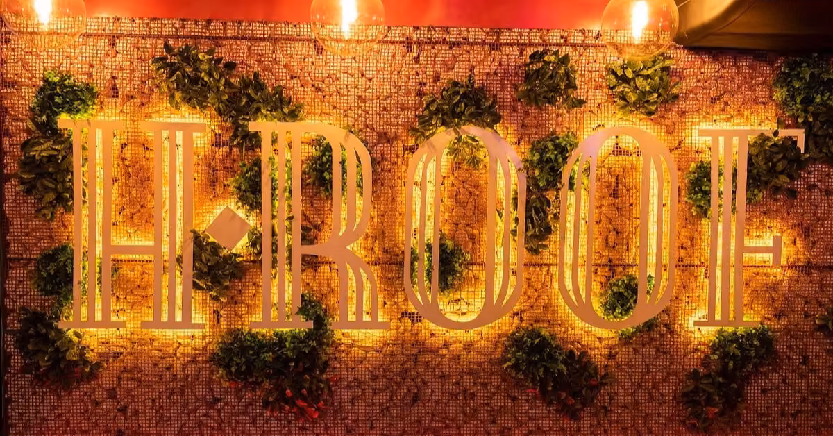 Illuminated H-ROOF sign on a tiled wall with green plants and yellow lights, creating a vibrant, inviting nightlife atmosphere.