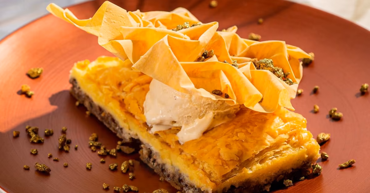 Baklava slice topped with ice cream and crisp phyllo, sprinkled with pistachios on a terracotta plate.