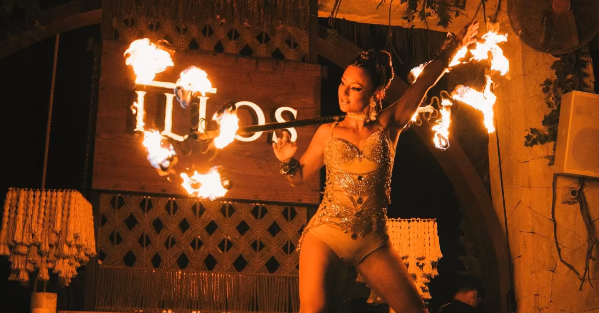 Performer in a sparkling costume twirls a fire baton, flames arching before a wooden ILIOS sign at a dimly lit restaurant.