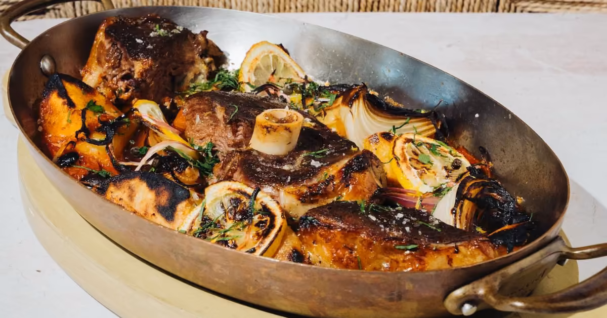 A roasting pan with browned meat, lemon slices, onions, and herbs, all golden and perfectly roasted at Ilios Restaurant.