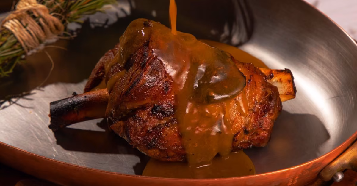 Roasted meat shank in a copper pan at Ilios Restaurant, topped with brown gravy; herbs rest in the background.