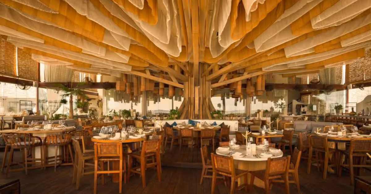 Ilios Restaurant features wooden furniture, round dining tables, a central tree structure, and warm fabric draped from the ceiling.