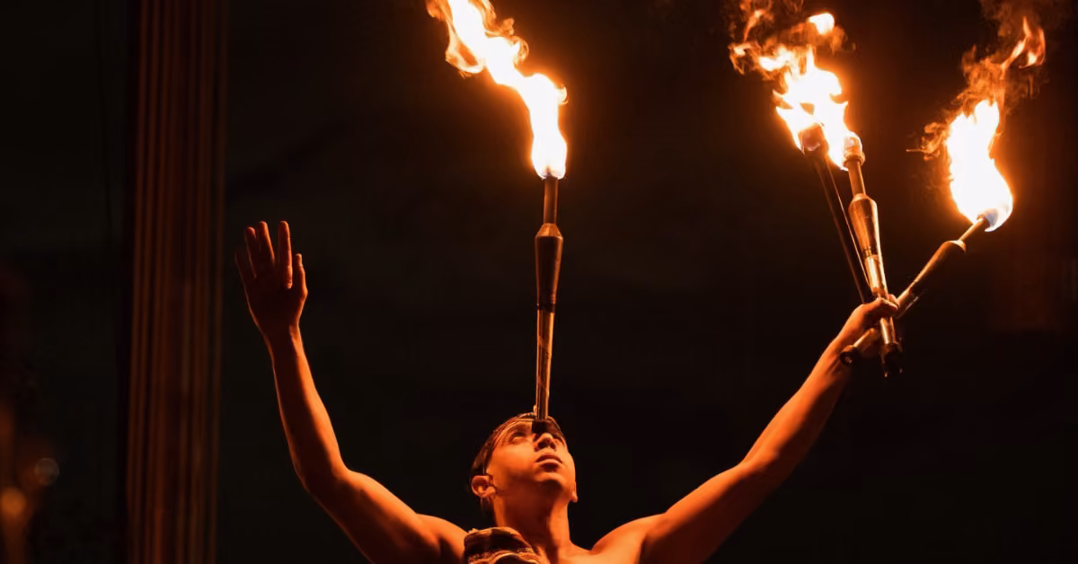 A performer balances a flaming torch on their head and holds two more, one in each hand, against a dark background.