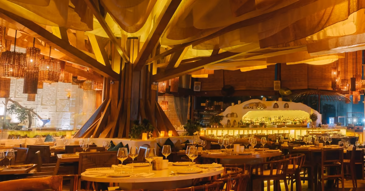 Ilios Restaurant with a large tree centerpiece, round tables, wooden chairs, and warm ambient lighting for a cozy atmosphere.