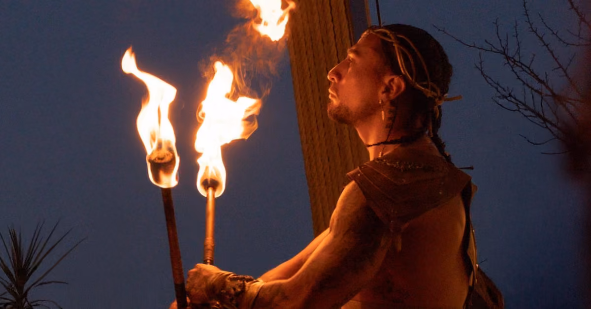 A tattooed performer with braided hair holds two flaming torches outside Ilios Restaurant at dusk, plants and a tree behind them.