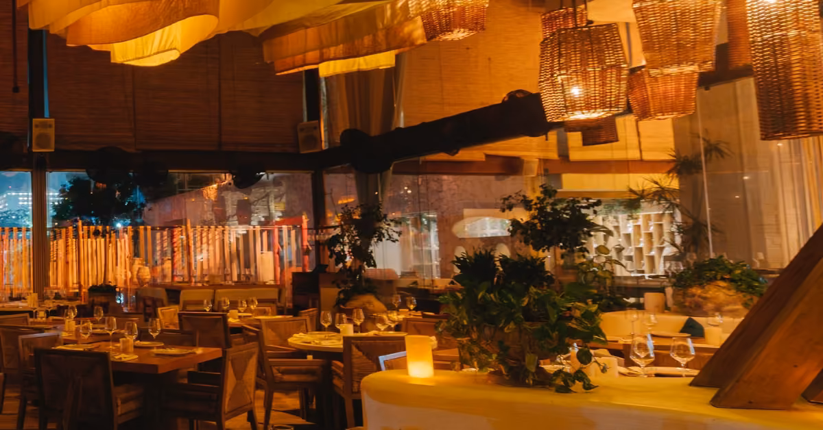 Cozy restaurant with warm lighting, wooden tables, glassware, woven lamps, potted plants, and large windows.