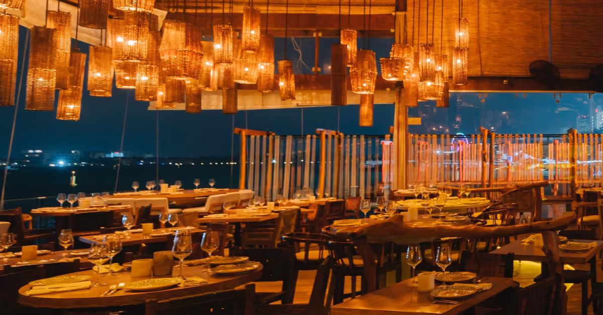 Ilios Restaurant at night with cozy lighting, wooden tables, woven pendant lights, and large windows overlooking the city.