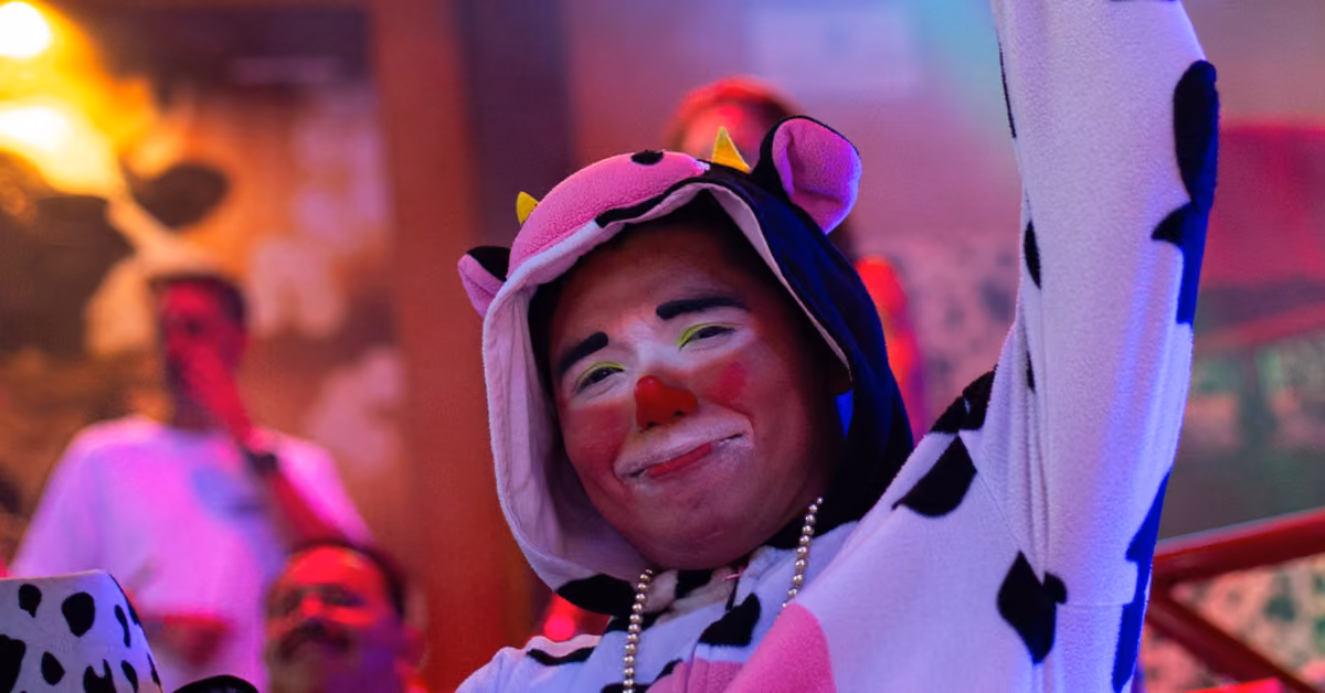 A person in a cow costume with bright face paint smiles and raises an arm at a lively, colorful night club with blurred guests.