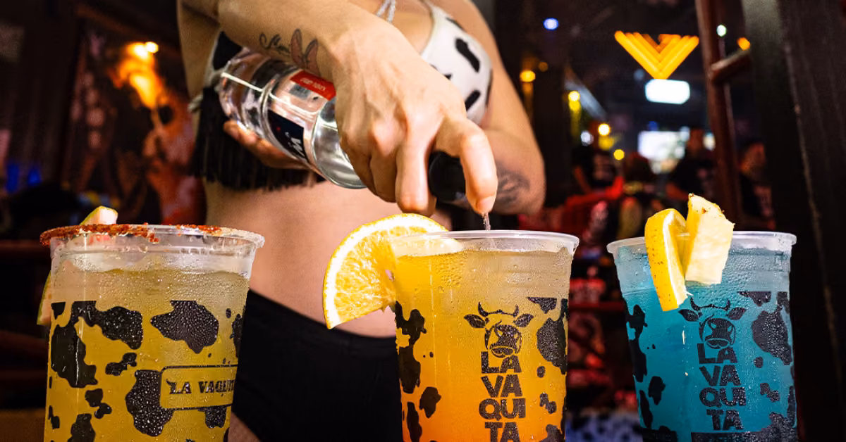 Someone pours a clear drink into a cup with an orange slice. Two bright, citrus-garnished drinks sit in La Vaquita cups nearby.