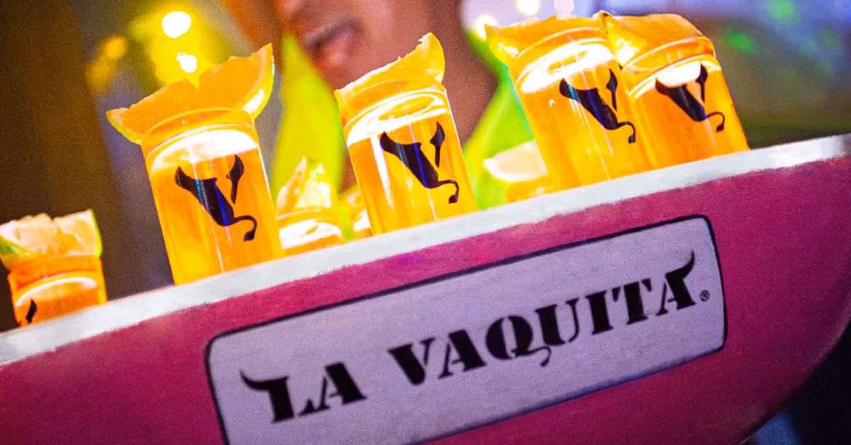Orange drinks with orange slices in clear cups, black cow logo, sit in a pink LA VAQUITA tray; blurred person behind.