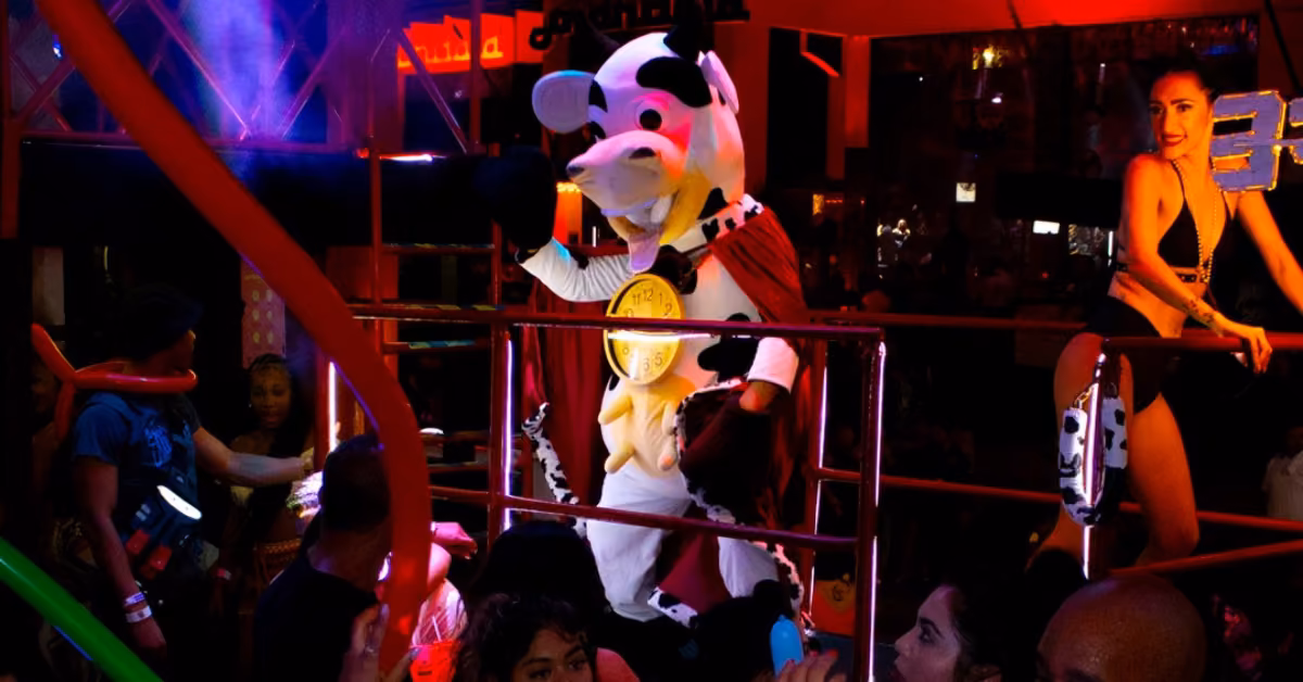 A person in a cow costume with a cape stands on stage at La Vaquita, surrounded by dancing people and colorful party lights.