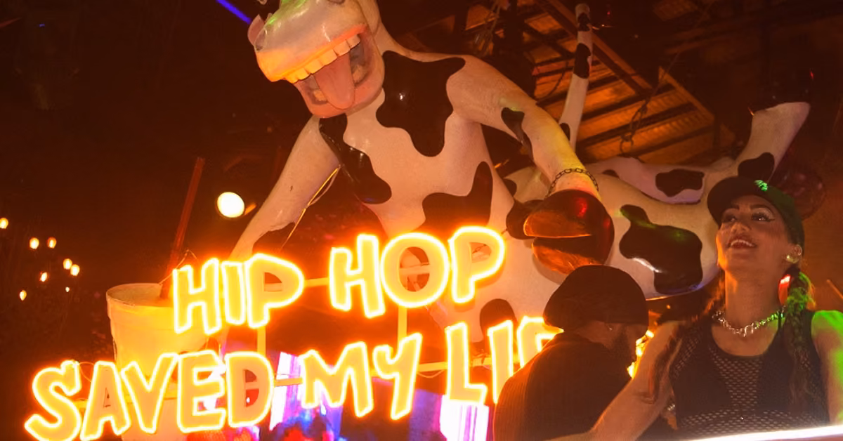 A cartoon cow figure hangs above neon orange “HIP HOP SAVED MY LIFE” text in La Vaquita Night Club with colorful lights.