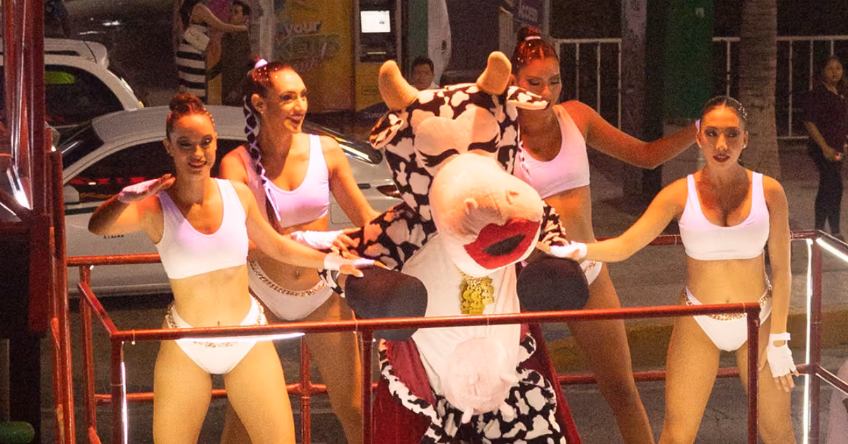 A person in a cow costume dances with four women in white outfits on a bright stage at La Vaquita, with people in the background.