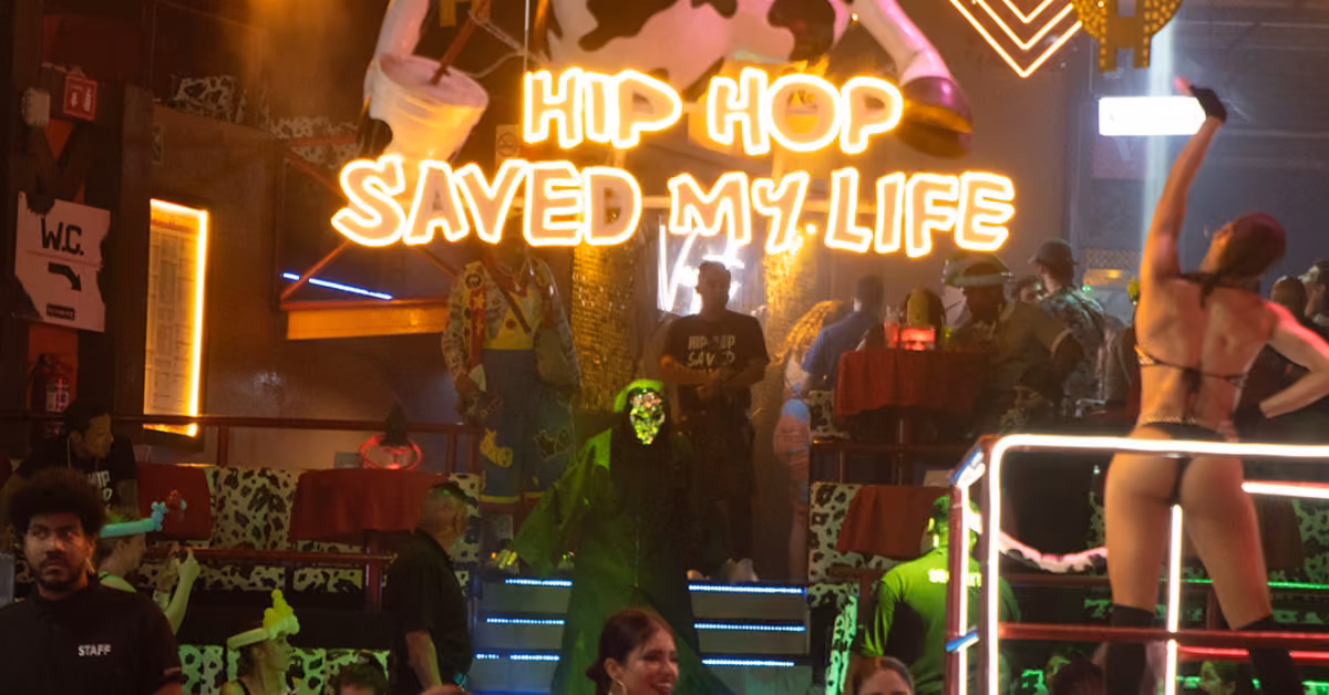 Crowded night club with neon HIP HOP SAVED MY LIFE sign, dancers, a woman posing on a platform, and cow-pattern decor.