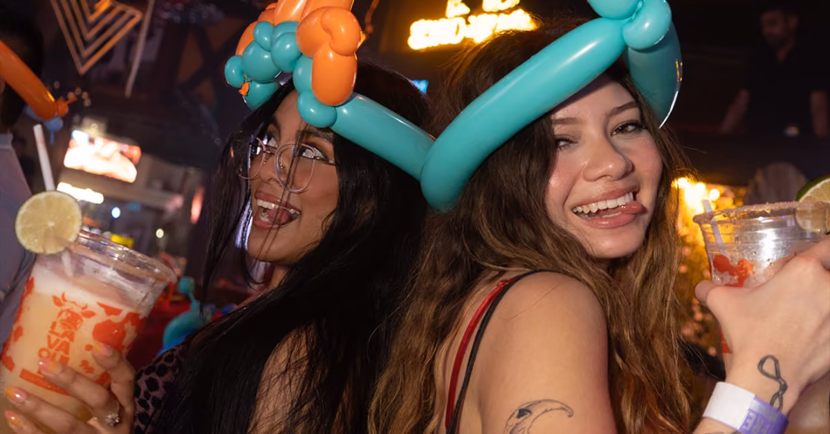 Two women with balloon hats smile at a colorful party, holding lime drinks; one playfully sticks her tongue out.