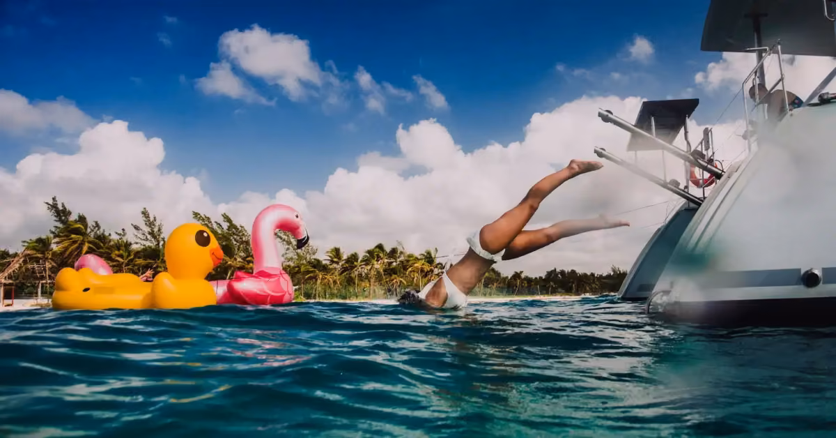 A person in a white swimsuit dives off a cruise boat into blue water near big inflatable duck and flamingo floats, with palms behind.