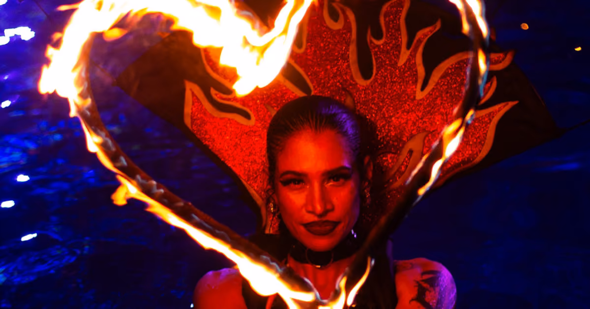 A woman in bold makeup and a fiery red headdress stands before a glowing heart of flames at Mandala Beach Club, blue backdrop.