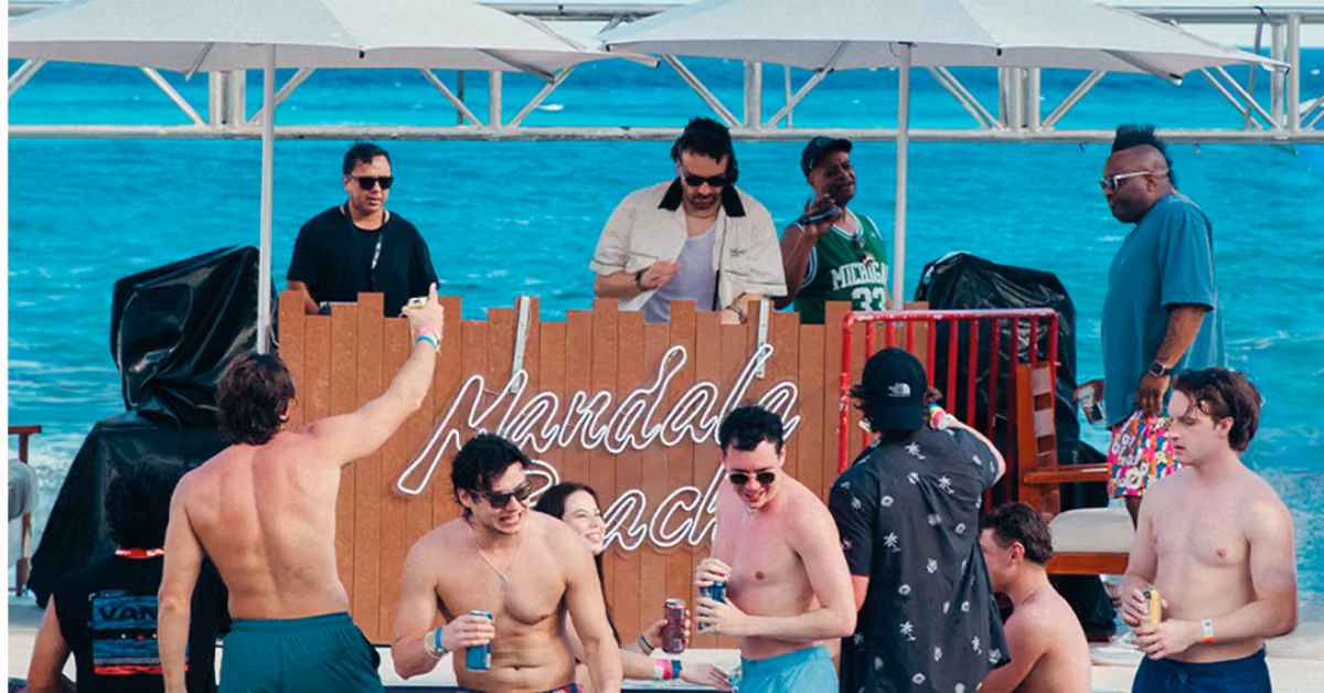 Friends in swimwear relax with drinks at Mandala Beach Club’s outdoor bar as a DJ plays and some guests dance by the sea.