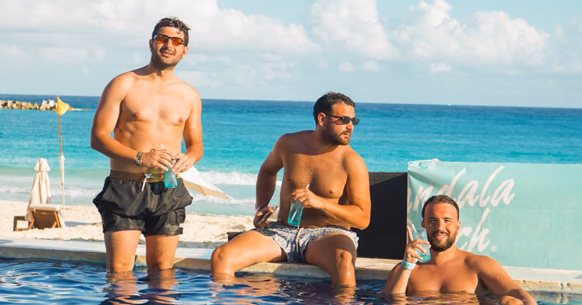 Three men in swim trunks relax by the pool with drinks, a sandy beach and clear blue ocean behind them under a sunny sky.