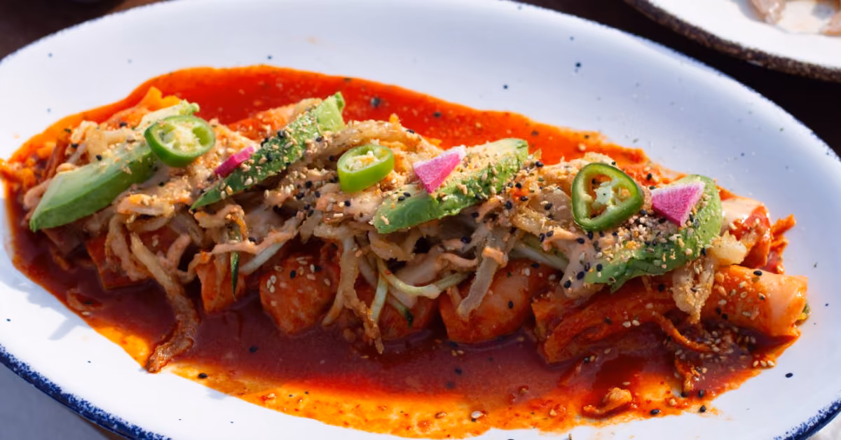 Kimchi with sliced avocado, green chili, shredded veggies, sesame seeds, and purple radish in a vibrant red sauce.