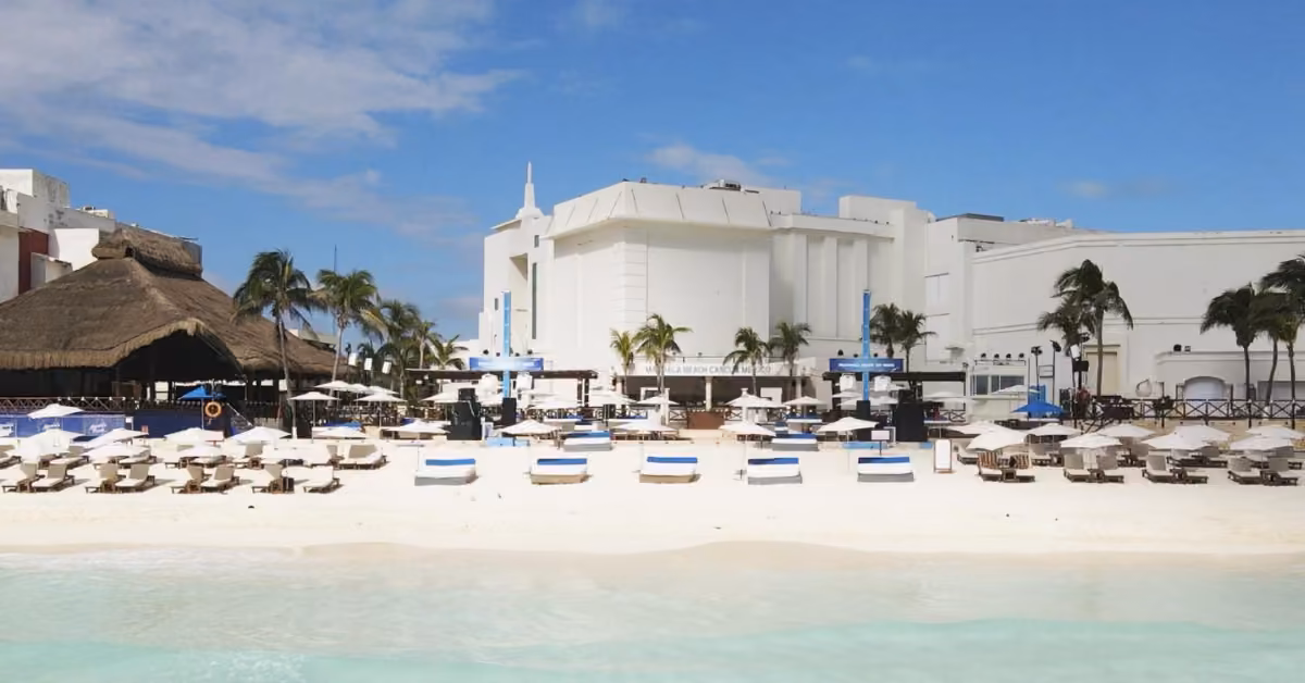 White sand beach with lounge chairs, umbrellas, palm trees, and nearby white and thatched-roof buildings under blue skies.