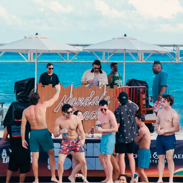 People in swimsuits dance, mingle, and enjoy drinks at a lively beach party by the ocean as a DJ plays music under umbrellas.