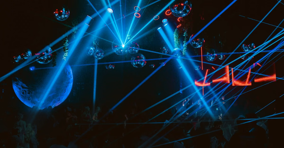 A dimly lit night club with blue laser lights, disco balls, and a dancing crowd. Red neon letters and a moon hang in back.