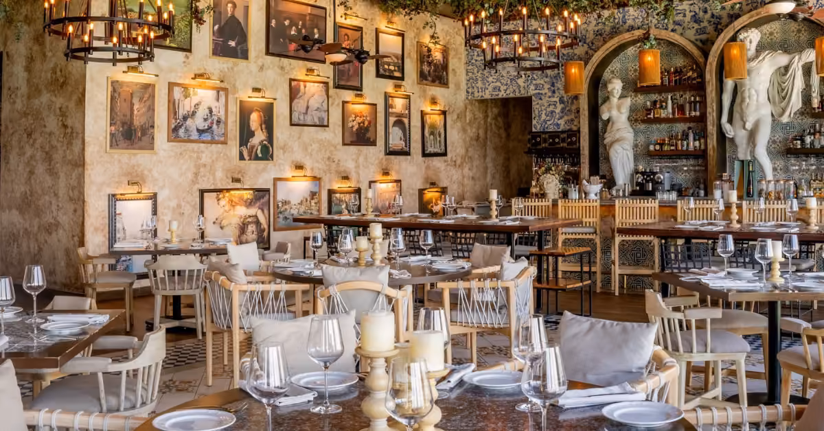 Nicoletta restaurant with wooden chairs, set tables, chandeliers, candles, framed art, greenery, and two white statues by the bar.