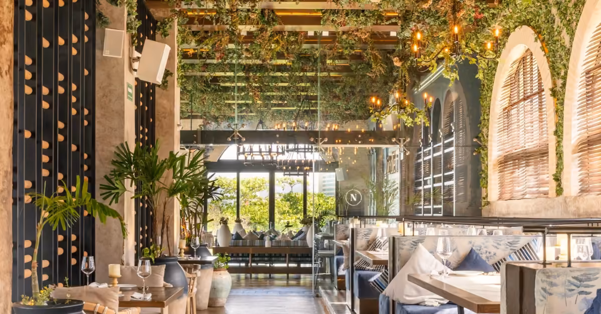 Nicoletta Restaurant’s chic interior has blue and white booths, tall plants, big windows, and greenery hanging from the ceiling.