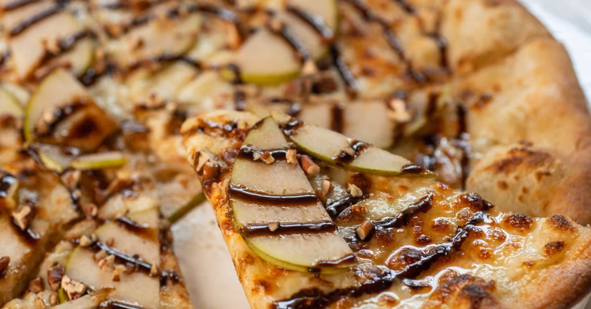 Pizza topped with pears, nuts, melted cheese, and balsamic glaze, with one slice being pulled from the pie.