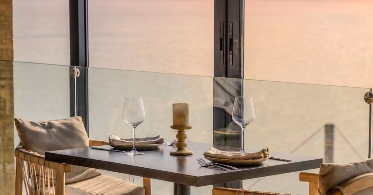 Table for two with wine glasses and candle by a window, overlooking the lagoon at sunset. Cozy chairs face each other.