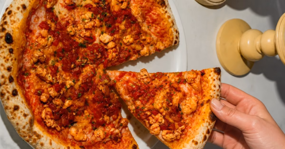 A hand lifts a sausage-topped pizza slice from a whole pie on a white plate, with a yellow candle holder in the background.