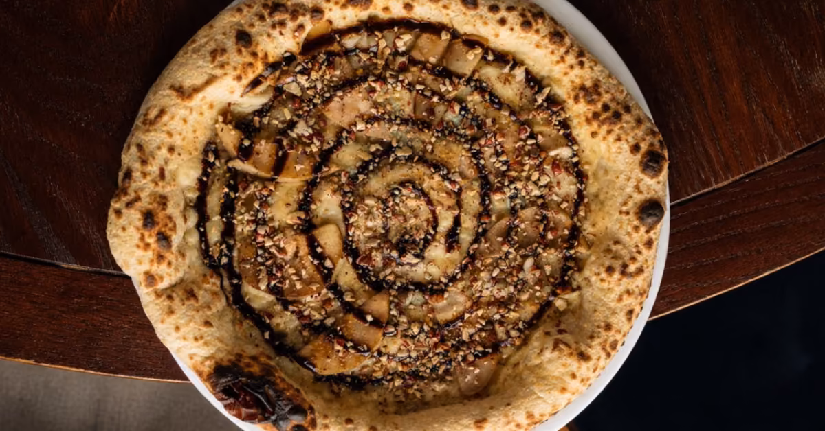 Pizza with thick crust, dark spiral sauce, nuts, and fruit on a white plate at Nicoletta Restaurant’s dark wood table.