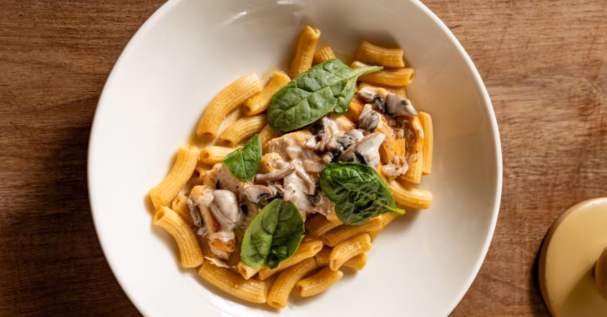 Rigatoni pasta with creamy mushroom sauce and fresh spinach leaves, served in a bowl on a wooden table.