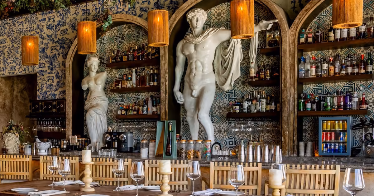 Bar at Nicoletta with classical statues, bottles, wicker chairs, hanging lamps, blue tiles, greenery, and a set wooden table.