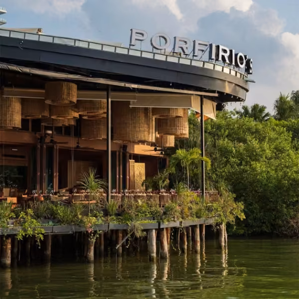 Porfirio’s sits on stilts above the water, surrounded by greenery and hanging lights, with tables under a partly cloudy sky.