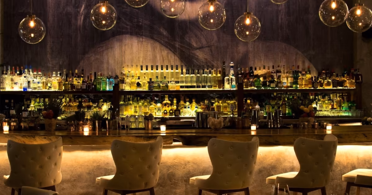 Warmly lit bar at Rosa Negra with shelves of liquor, pendant lamps, marble counter, candles, and five cushioned barstools.