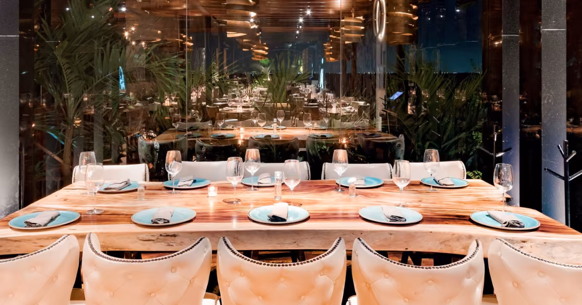 Elegant restaurant with a wooden table for eight, white chairs, blue plates, black napkins, soft lighting, and green plants.